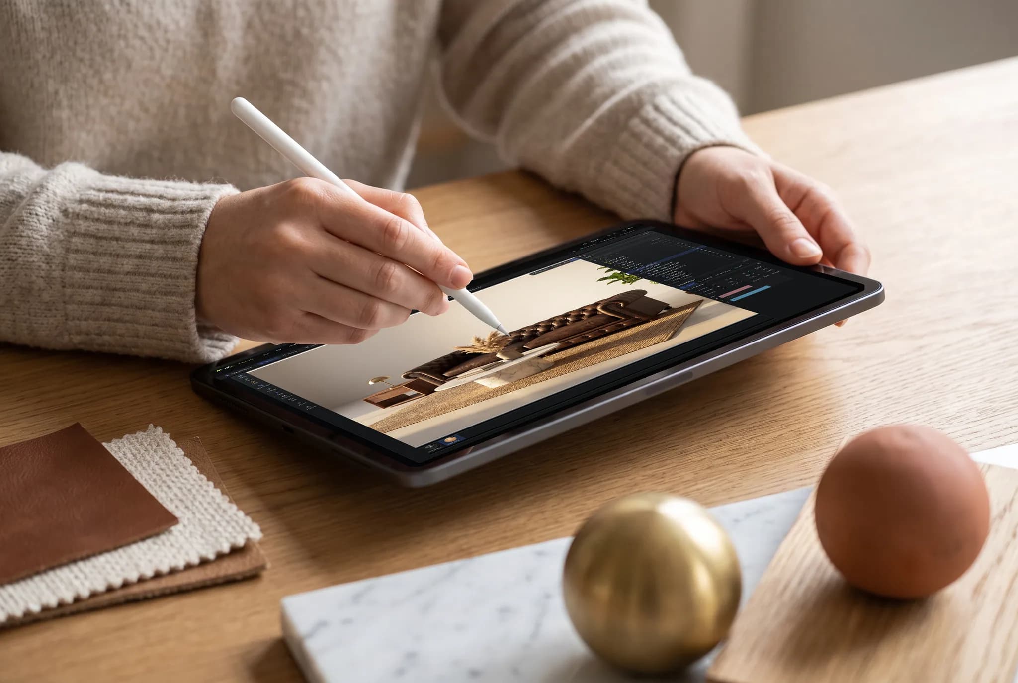 Two hands holding an iPad running Studio, an Apple Pencil touching the screen, with a desk scene rendered in the viewport.